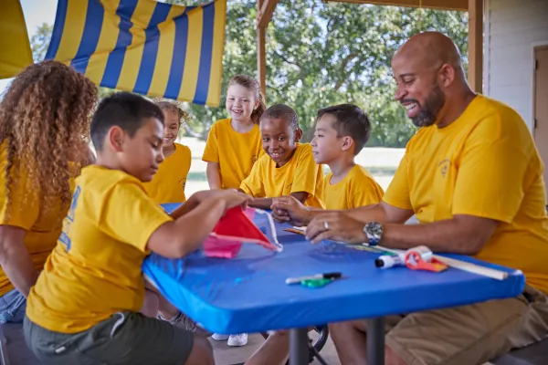 An adult leader works alongside Cub Scouts building kites at a picnic table during day camp.