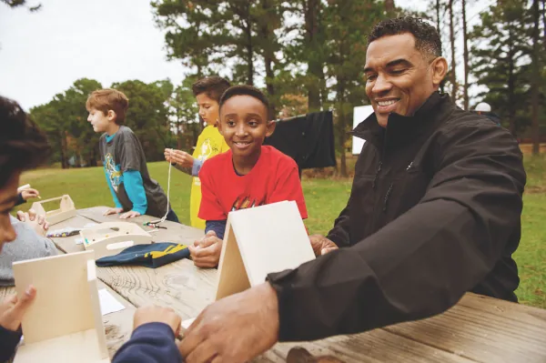 A smiling Cub Scout leader helps Scouts assemble wooden birdhouses at a picnic table during an outdoor pack activity.