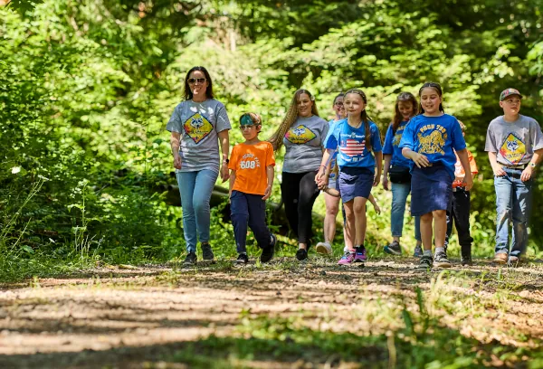 Cub Scouts and adult den leaders hiking together along a sunlit forest trail on a summer pack outing.