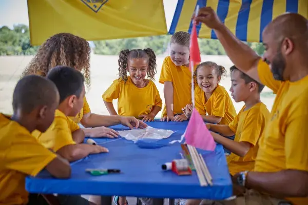 Six Cub Scouts and two adult leaders build kites together at an outdoor pack activity under a blue and gold canopy.